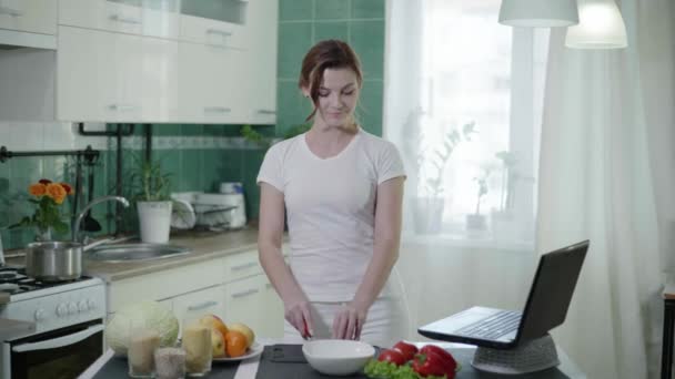 Mädchen in lässiger Hauskleidung bereitet in der Küche am Tisch einen gesunden Salat zu, schneidet mit dem Messer Gurken und blickt auf den Laptop-Bildschirm — Stockvideo