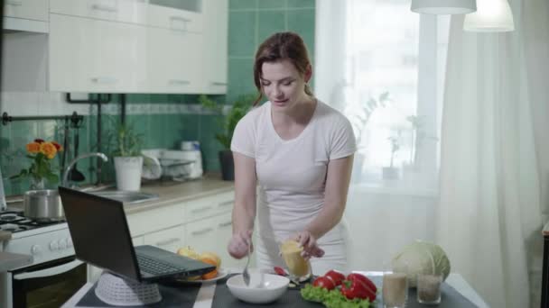 Gesunde Ernährung, Mädchen in lässiger Kleidung misst einen Weizenbrei mit Löffel während der Zubereitung von Salat und schaut Laptop auf Bildschirm am Tisch in der Küche — Stockvideo