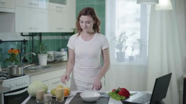 Домохозяйка использует ноутбук во время приготовления здоровой пищи на кухне, девушка измеряет кашу пшеницы с ложкой и смотреть рецепт на экран — стоковое видео