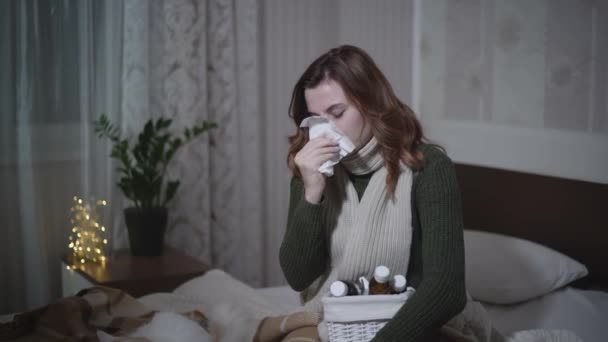 Beteg nő meleg sálban szenved a hőmérséklet és a fejfájás alatt influenza ül az ágyon zsebkendővel és gyógyszerek privát apartman — Stock videók