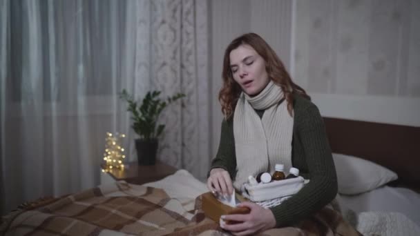 Nemocná dívka v teplém oblečení s ucpaným nosem kýchá a kašle, zatímco sedí na posteli s ubrousky a léky, nemocná žena používá kapky z nemoci — Stock video