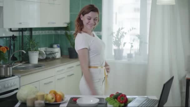 Portret van meisje na een succesvol dieet meet haar taille met een meetlint en poseren in de keuken met een appel en peper in haar handen — Stockvideo