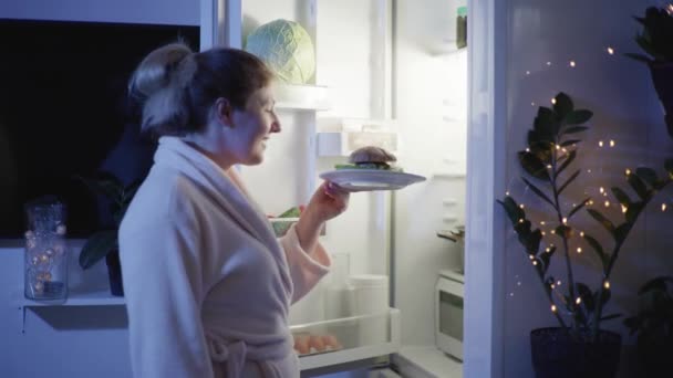 Upset woman on diet, hungry girl opens the refrigerator at night and happily get a plate with a gaburger then sadly takes glass and pours water for drinking — Stock Video