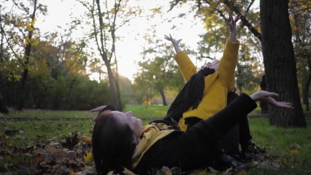 Stagione autunnale, madre felice con una figlia carina sdraiata sul prato nel parco autunnale e gettare foglie durante una passeggiata in giardino — Video Stock