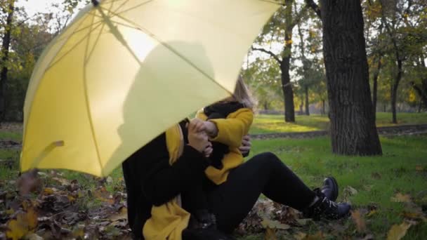 Sevgi dolu anne parkta güzel kızıyla oynuyor. Sonbahar parkında çimlerde oturuyor. Şemsiyenin altında dağınık yapraklar var. — Stok video