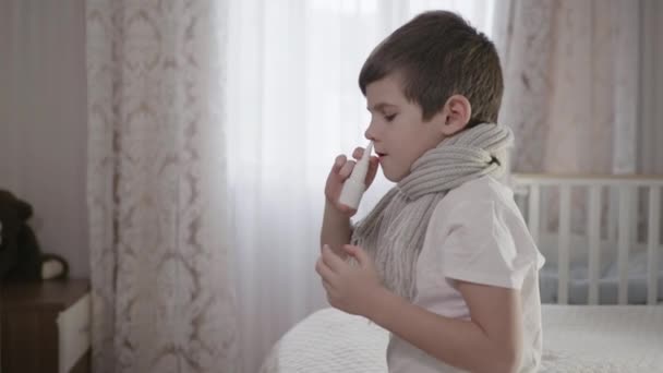 Enfant malade garçon dans une écharpe chaude avec nez bouché utilise des gouttes de la maladie pendant la grippe assis sur le lit dans un appartement privé — Video