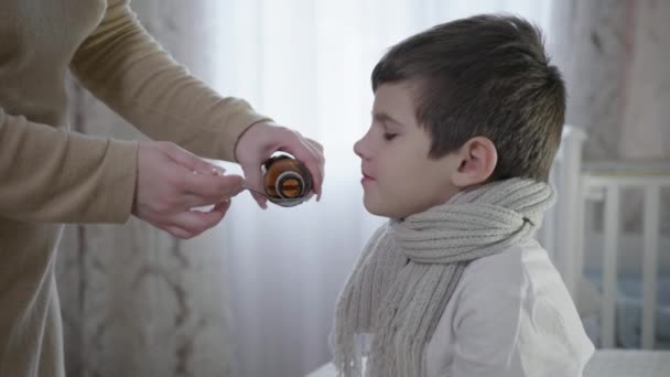 Soin des enfants, parent verse du sirop contre la toux dans une cuillère pour enfant malade prend des médicaments pour la maladie assis sur le lit dans une écharpe chaude — Video