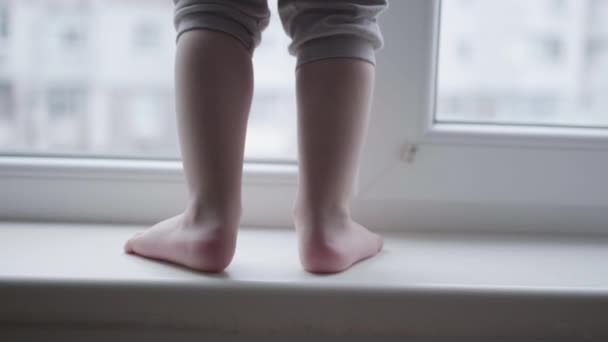 Детские ноги на подоконнике, маленький мальчик в опасности подходит к окну близко — стоковое видео
