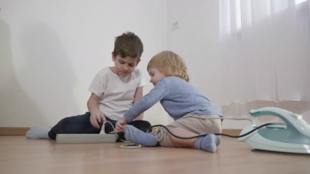 Дети играют опасно с электрическим удлинителем шнур, маленькие мальчики подключить провода вилки из железа на пол в комнате — стоковое видео