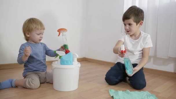 Niños juegan peligrosamente con productos químicos domésticos, niño pequeño con detergente spray botella en un trapo sentado en el suelo junto a su hermano menor que tira guantes — Vídeo de stock