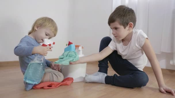 Livshotande, liten pojke spray hushållskemikalier från en plastflaska på en trasa som innehas av en äldre bror sitter på golvet i rummet — Stockvideo