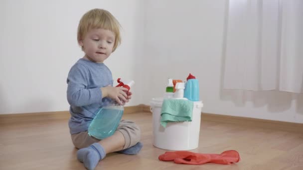 Παιδί σε κίνδυνο κάθεται κοντά σε ένα κουβά με πλαστικά μπουκάλια με χημικά οικιακής χρήσης, μικρό αγόρι ψεκάζει γυαλί απορρυπαντικό στο γάντι καουτσούκ στο πάτωμα στο δωμάτιο — Αρχείο Βίντεο