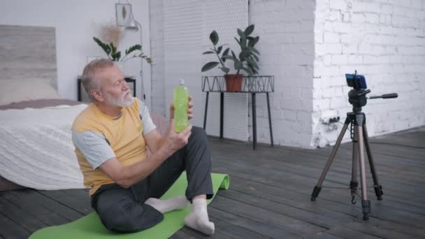 Influential elderly male blogger records video review of vitamin water for blog on a smartphone recommending his subscribers drink for sports sitting on yoga mat in beautiful interior — Stock Video