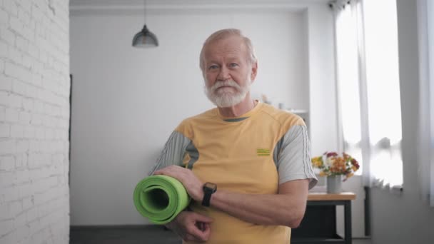 Portret van een knappe oudere man met een yogamat in zijn handen en een slim horloge na het sporten om zijn gezondheid staande te houden in de ruimte tegen een prachtig interieur — Stockvideo