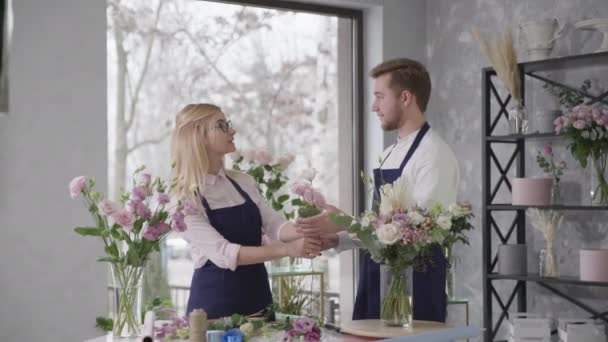 Floristas de trabalho em equipe, jovens profissionais sorrindo loira e encantador menino coletar composição de flores na loja moderna vendendo flor — Vídeo de Stock