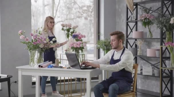 花店里，专业花店里的女士和男士请网上商店点菜，让他们把花束放在桌旁，并带着笔记本电脑 — 图库视频影像