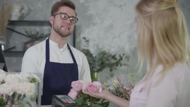Vonzó fiatal férfi eladók dolgozik virágüzlet pénztár, érintésmentes fizetést terminálon keresztül mobiltelefon az ügyfél fizet csokor gyönyörű virágok, sikeres üzlet — Stock videók