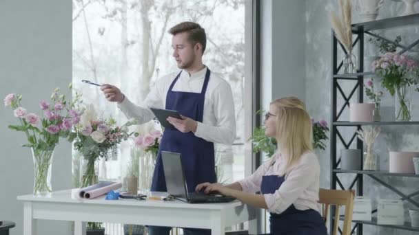 Молодая счастливая женщина работает за ноутбуком вместе с мужчиной флорист использует онлайн-сервис для приема заказов на доставку букета цветов из цветочного магазина, молодые предприниматели — стоковое видео