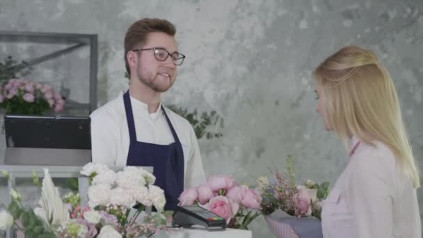 Ritratto di giovane uomo attraente, rappresentante della parità di genere, lavora come fiorista nel moderno negozio di fiori, vende bouquet di bei fiori a un cliente soddisfatto che paga per l'acquisto in contanti — Video Stock