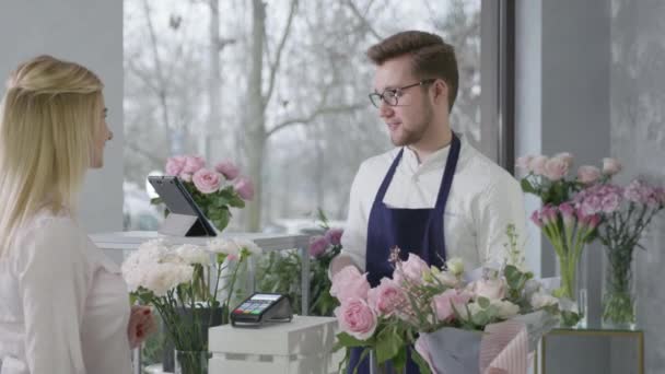 Bezdotykowe płatności, kobieta kupująca płaci bukiet kwiatów za pomocą smartfona w terminalu, młody mężczyzna sprzedawca kwiaciarnia przedstawiciel równouprawnienia płci pracuje przy kasie w butiku kwiatów — Wideo stockowe