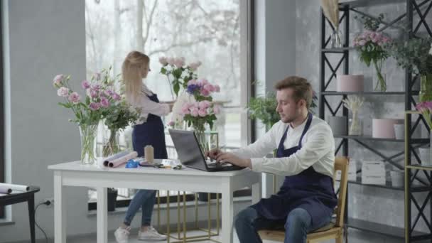 Доставка цветов, молодые современные профессиональные флористы работают в цветочном магазине и принимают заказы по телефону, сидя за столом с ноутбуком — стоковое видео