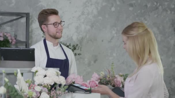 Portrait de jeune fleuriste homme, vendeur masculin vend bouquet de fleurs à une femme blonde acheteur, femme paie l'achat à la boutique de fleurs sur une base régulière par paiement sans contact en utilisant smartphone — Video
