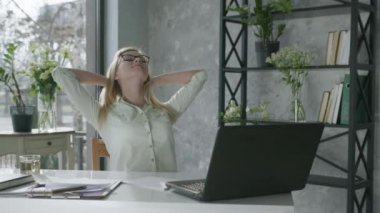 Serbest çalışan mutlu bir sarışının portresi, bilgisayar ve ofis bilgisayarlarıyla çevrimiçi çalıştıktan sonra rahatlamayı seviyor.