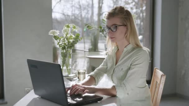 Werk online, mooie aantrekkelijke blonde vrouw werkt op laptop in bril voor visie en neemt notities in notebook, dan neemt af bril kijken naar camera en glimlachen, concept van succes — Stockvideo