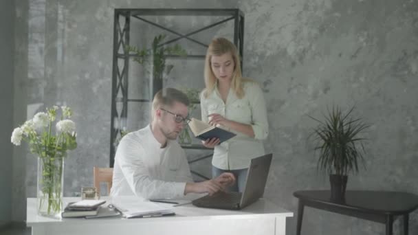 Stage, een jonge zakenvrouw met een boek in handen leert een stagiaire zitten op laptop, werknemer relaties — Stockvideo