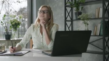 Bilgisayarda çalışan mutlu, güler yüzlü bir kadın taze çiçeklerle ofiste çalışıyor, başarıya seviniyor genç iş kadını online hizmet için dizüstü bilgisayar kullanıyor, siparişleri kontrol ediyor ve not defterine veri yazıyor.
