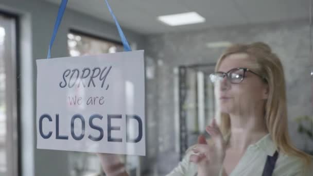 Propietario de un negocio una mujer atractiva en un delantal y gafas cambia el cartel en la puerta principal de CERRADO a ABIERTO, sonriendo a la exitosa apertura de una pequeña empresa — Vídeo de stock