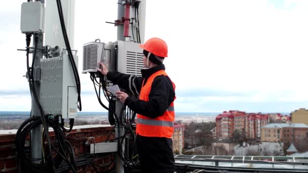 Pracownik techniczny mężczyzna w jasnej odzieży ochronnej pracuje na dachu w pobliżu anteny i robi notatki w notatniku — Wideo stockowe