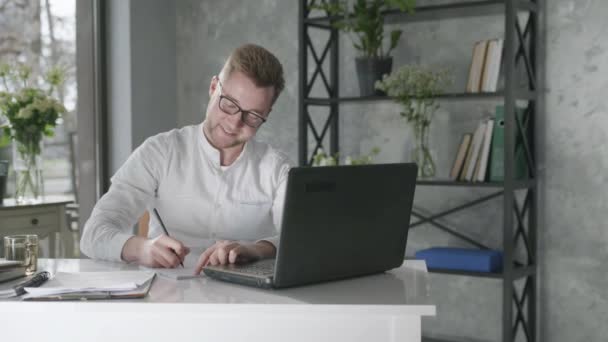 Fiatal férfi vállalkozó dolgozik laptop számítógép, miközben ül az asztalnál az irodában, örömteli munka jól végzett emelje fel a kezét fel a háttérben a friss virágok, koncepció a siker — Stock videók