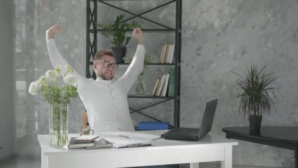 Радостный человек держит руки за голову, сидя на стуле рядом со столом с компьютером в офисе с современным цветочным интерьером на фоне свежих букетов цветов, счастливый работник мужчина — стоковое видео