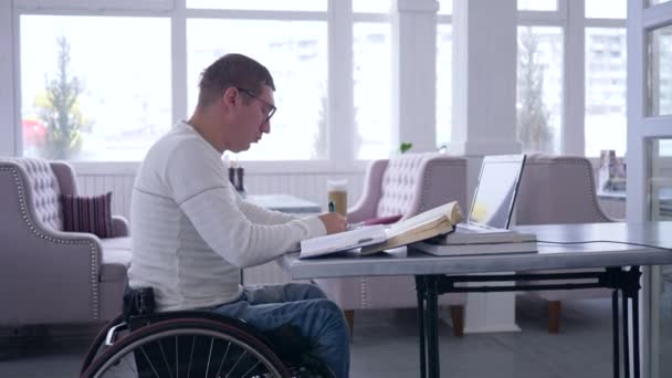 Business freelance, successful ailing male on wheel chair uses modern computer technology for remote work to development of business ideas and making notes in notebook close-up sitting at table — Stock Video