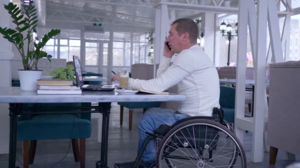 Handicapped man in wheel chair talking on mobile phone and working with a computer laptop sitting at table with cup of coffee in restaurant — Stockvideo