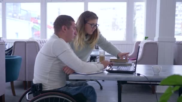 Individual lecture for disabled, successful educator female into eyeglasses conducts studying for invalid male on wheelchair using smart computer technology and books — Stockvideo