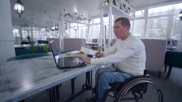 Educación en línea, estudiante discapacitado hombre en silla de ruedas utiliza la tecnología moderna portátil para aprender de lecciones en línea y libros haciendo notas en primer plano portátil sentado en la mesa en la cafetería — Vídeo de stock