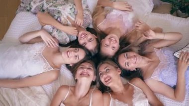 bachelorette party, gorgeous smiling friends girl in silk pajamas lie on bed and look into the camera in apartment