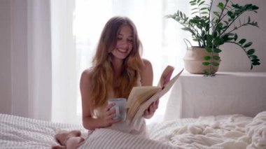 girl reads book with happy emotions and drink coffee at morning sitting on bed in light bedroom