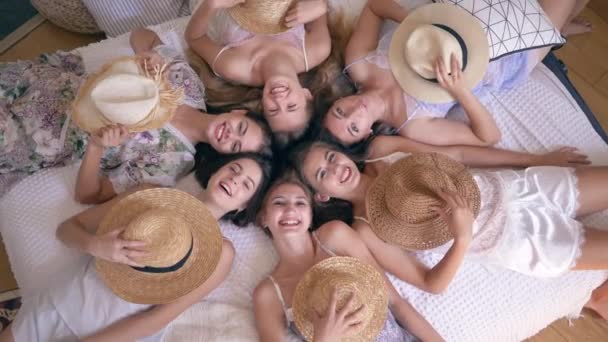 Joyful girlfriends in pajamas lie on bed with straw hats on faces and then take off them and smile into camera during hen party — Stockvideo