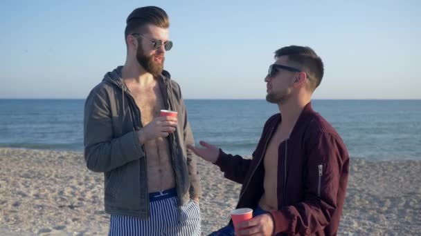 Male gossip, cheerful friends in glasses are drinking tea and talking on beach — ストック動画