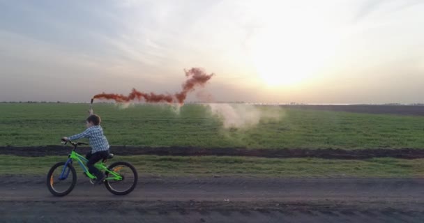 Criança aprende a andar de bicicleta, menino com uma bomba de fumaça colorida em uma estrada de campo — Vídeo de Stock