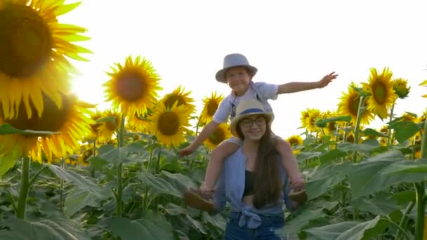 Vacaciones de verano del pueblo, chica adolescente lleva niño en el cuello y camina a través del campo con girasoles en la luz de fondo — Vídeo de stock