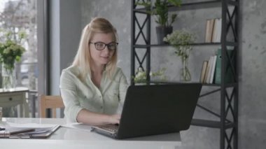 business online, happy attractive girl freelancer works at laptop computer in office with beautiful fresh flowers interior then shrugs hands to side rejoicing at job done and career success, plants