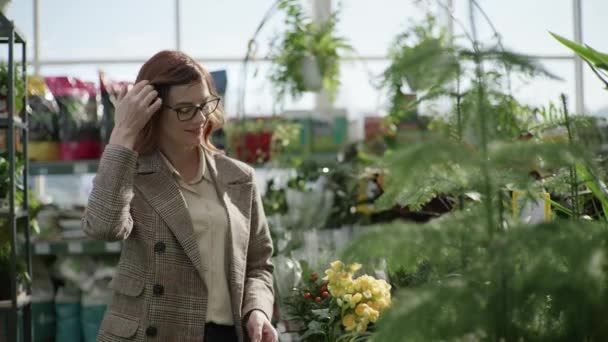 Portrait of a female florist in glasses for vision choosing decorative plants in pots for home or office design in flower shop standing on background of green plants, gardening — 图库视频影像