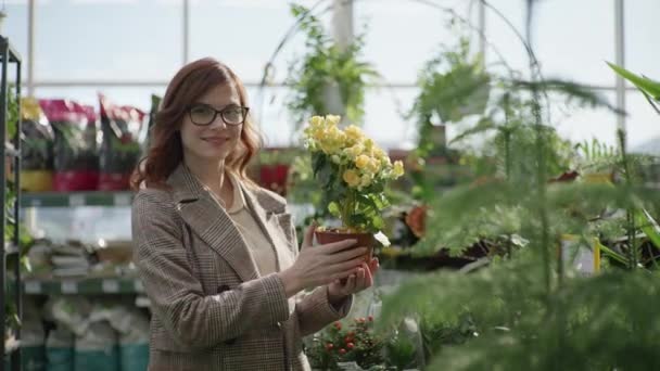 Portrait of female florist with glasses for vision choosing decorative blooming plants in pots for home or office design in flower shop standing on background of green plants, gardening — Stok video