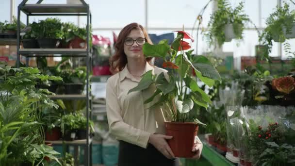 Portrait of young smiling girl with glasses for vision going away through greenhouse in flower shop with flowering ornamental plant in hands background of home plants — Stockvideo