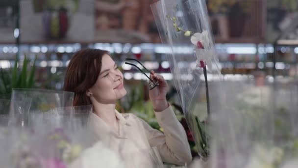 Atractivo feliz comprador femenino elige flor potted de la orquídea en invernadero de la tienda de flores en medio de plantas ornamentales verdes plantas — Vídeo de stock
