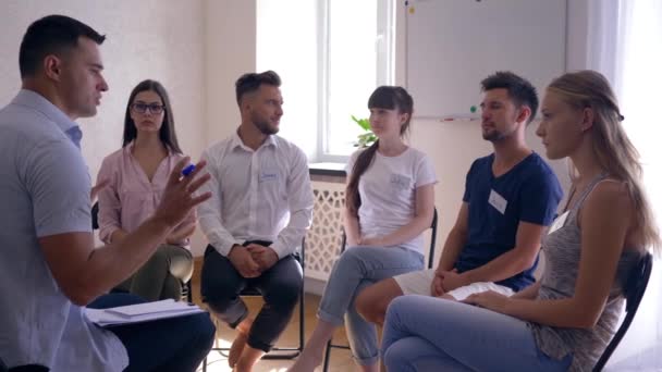 Групповая терапия, молодые люди со значками беседуют вместе с психологом, сидящим на стульях — стоковое видео